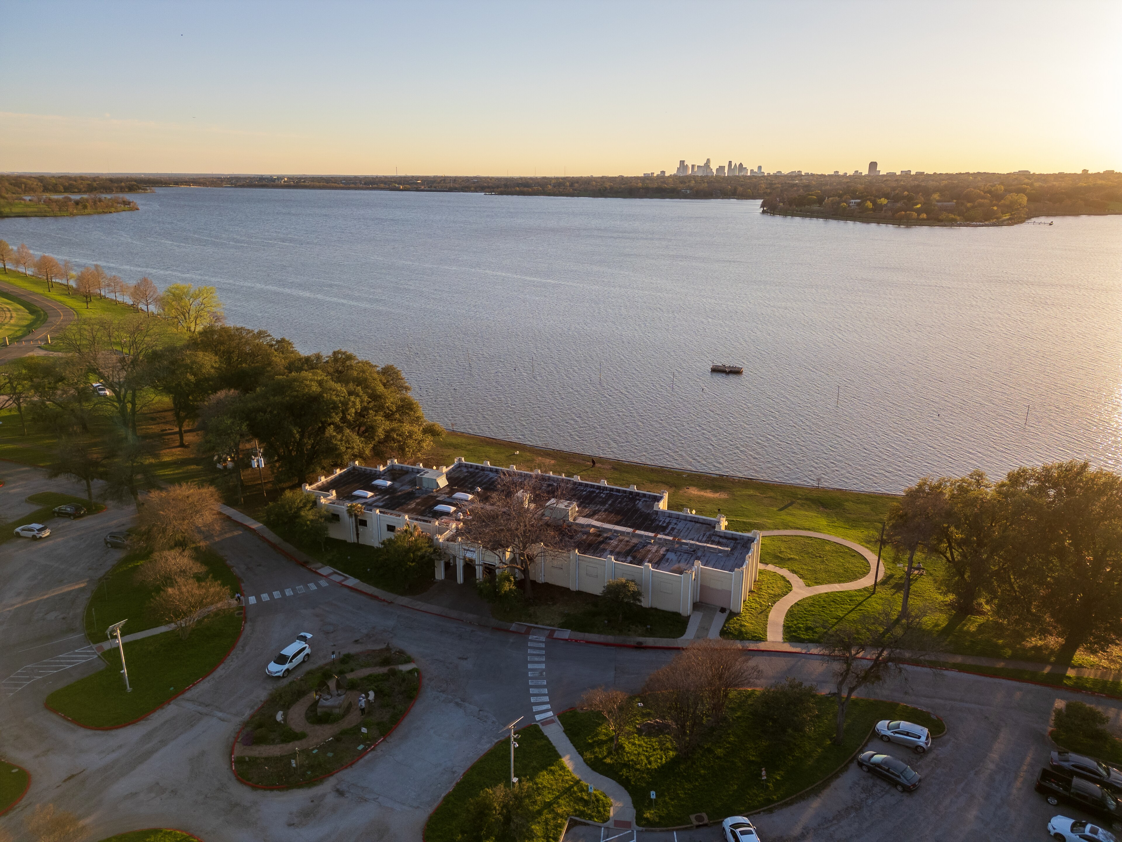 An aerial view of White Rock Lake at golden hour, with the low white Art Deco Bath House Cultural Center on the near shore and the distant downtown Dallas skyline on the horizon.