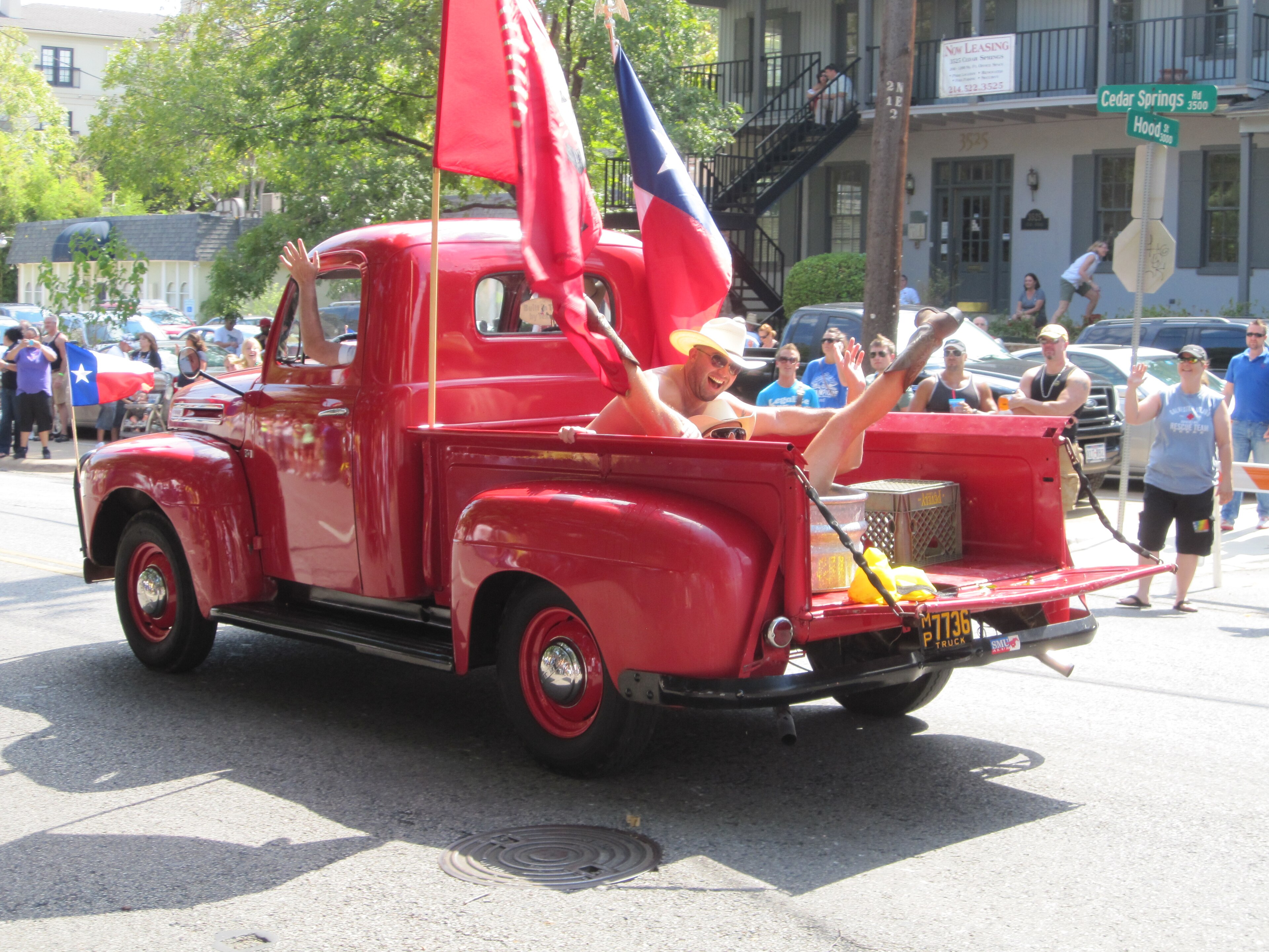 A vintage red pickup truck rolling down Cedar Springs Road during a Dallas Pride parade, a rider waving a Texas flag from the truck bed as onlookers line the sidewalk.