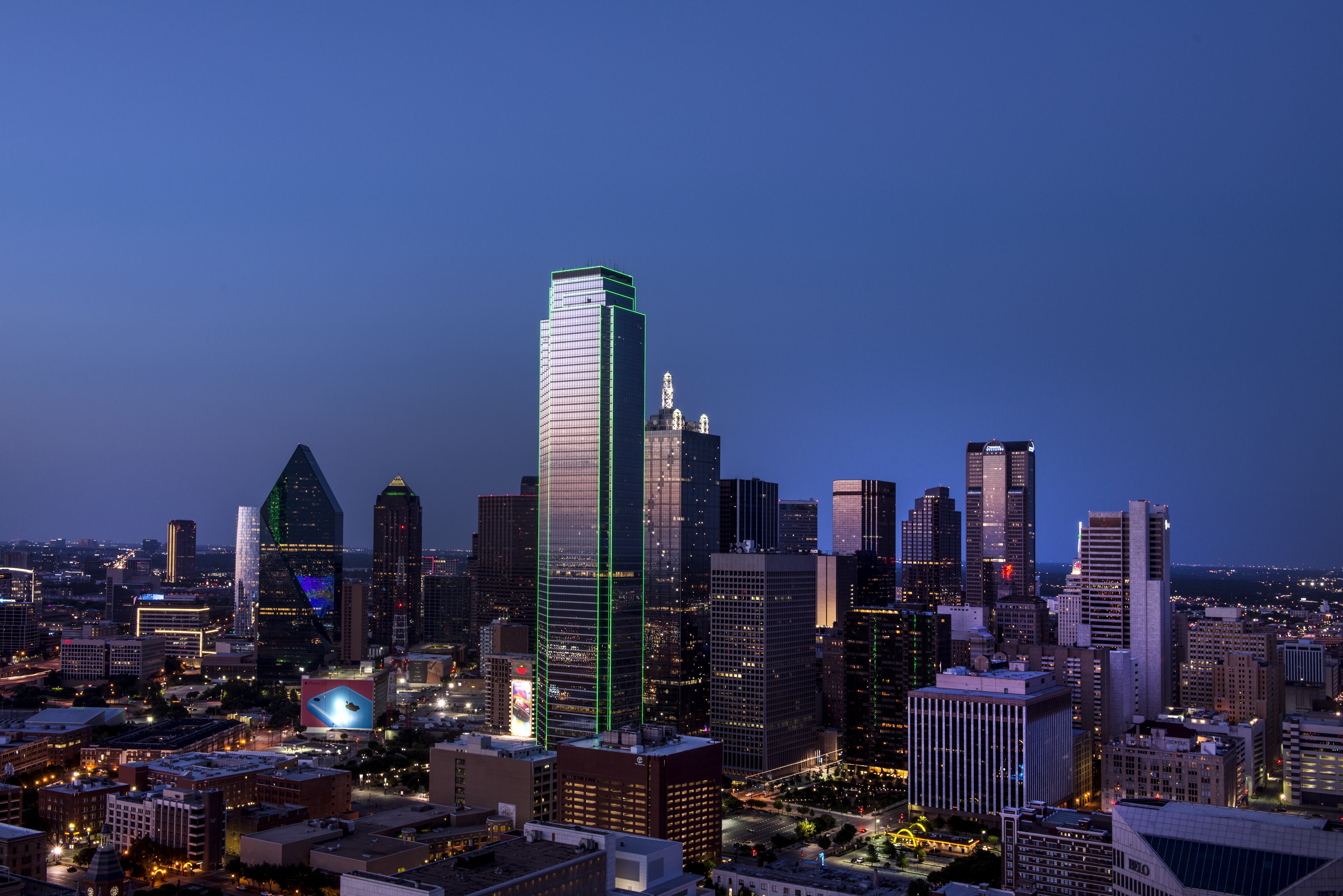 The downtown Dallas skyline at dusk under a deep blue sky, its tallest tower outlined in bright green light and the streets below lit amber.